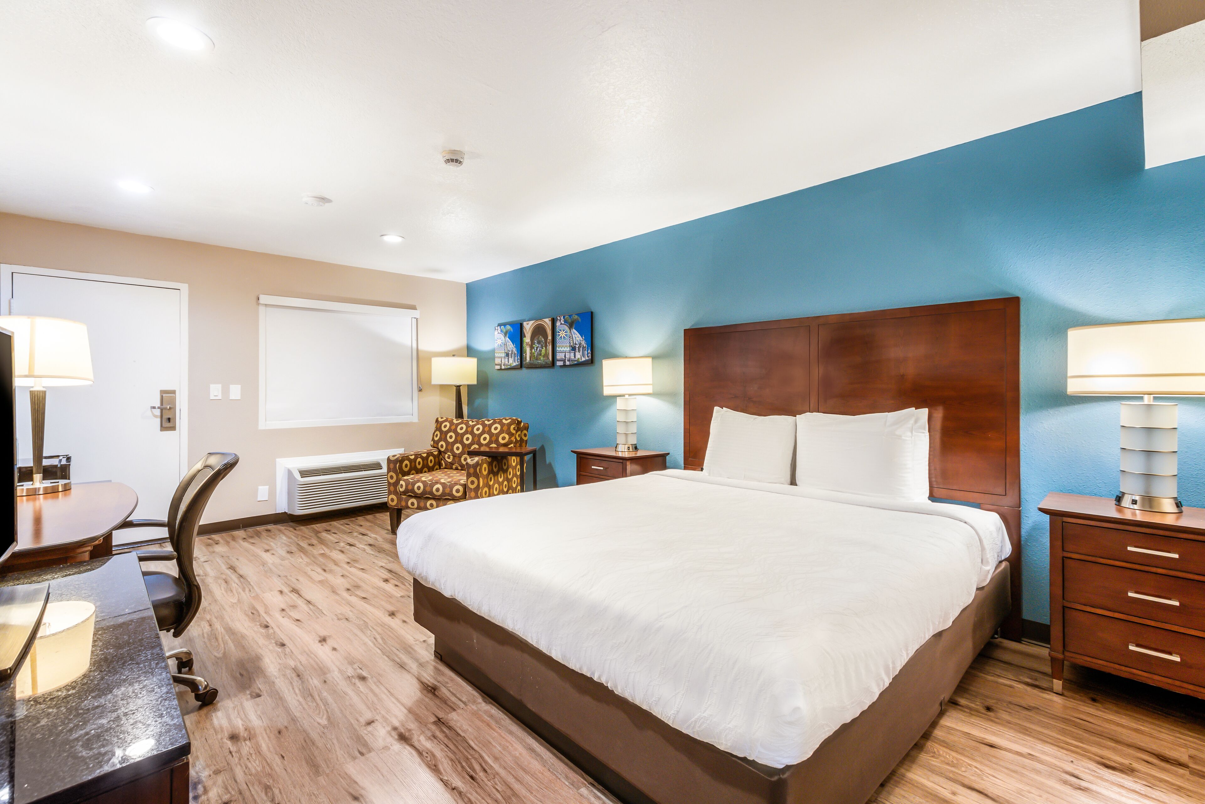 A hotel room with a large bed, a desk with a chair, a desk lamp, a wall-mounted heater, and a nightstand with a lamp.