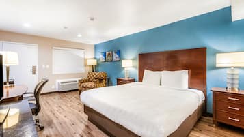 A hotel room with a large bed, a desk with a chair, a desk lamp, a wall-mounted heater, and a nightstand with a lamp.