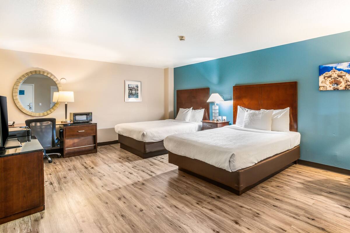 A hotel room with two beds, a desk, a chair, a desk lamp, a mirror, and a framed picture on the wall.