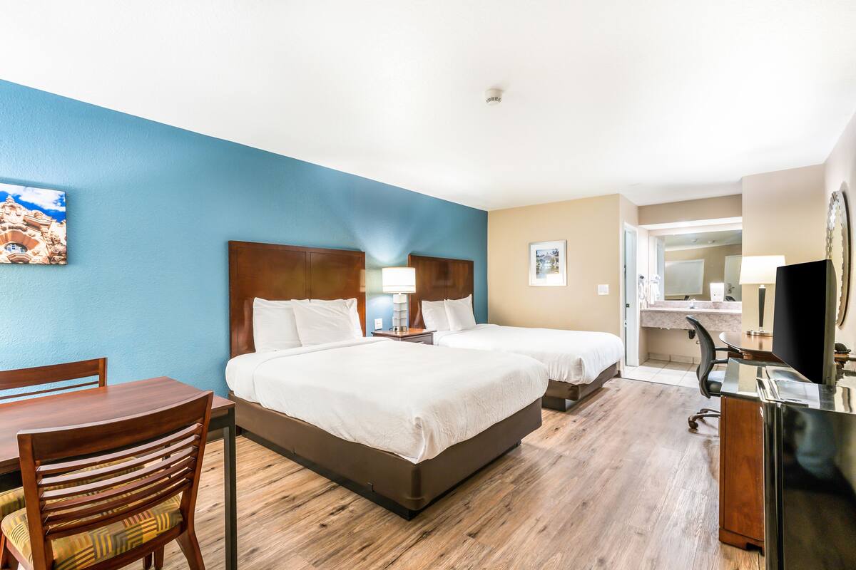 A hotel room with two beds, a desk, a chair, a TV, and a bathroom visible through an open door.