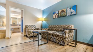 Una sala de estar con un sofá estampado, una mesita y una lámpara. Hay cuadros colgados en la pared y un acceso a otra habitación.