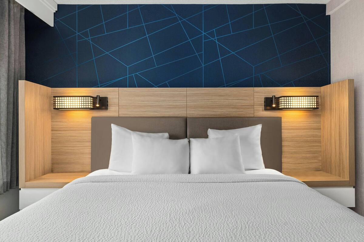 A modern hotel room with a large bed, wooden headboard, and geometric-patterned ceiling.