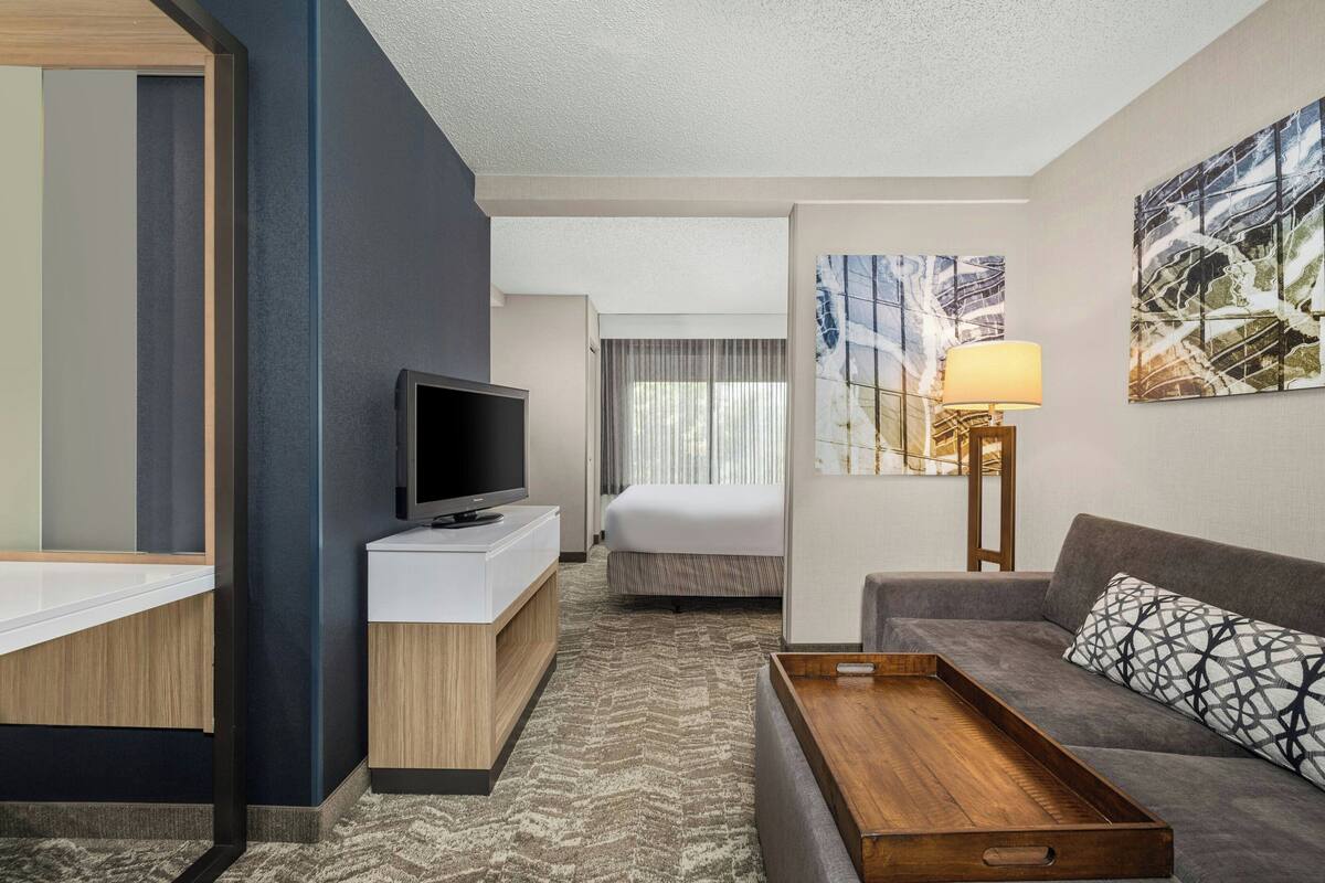 A modern hotel room with a flat-screen TV, a sofa, a bedside table, and a large window with curtains.