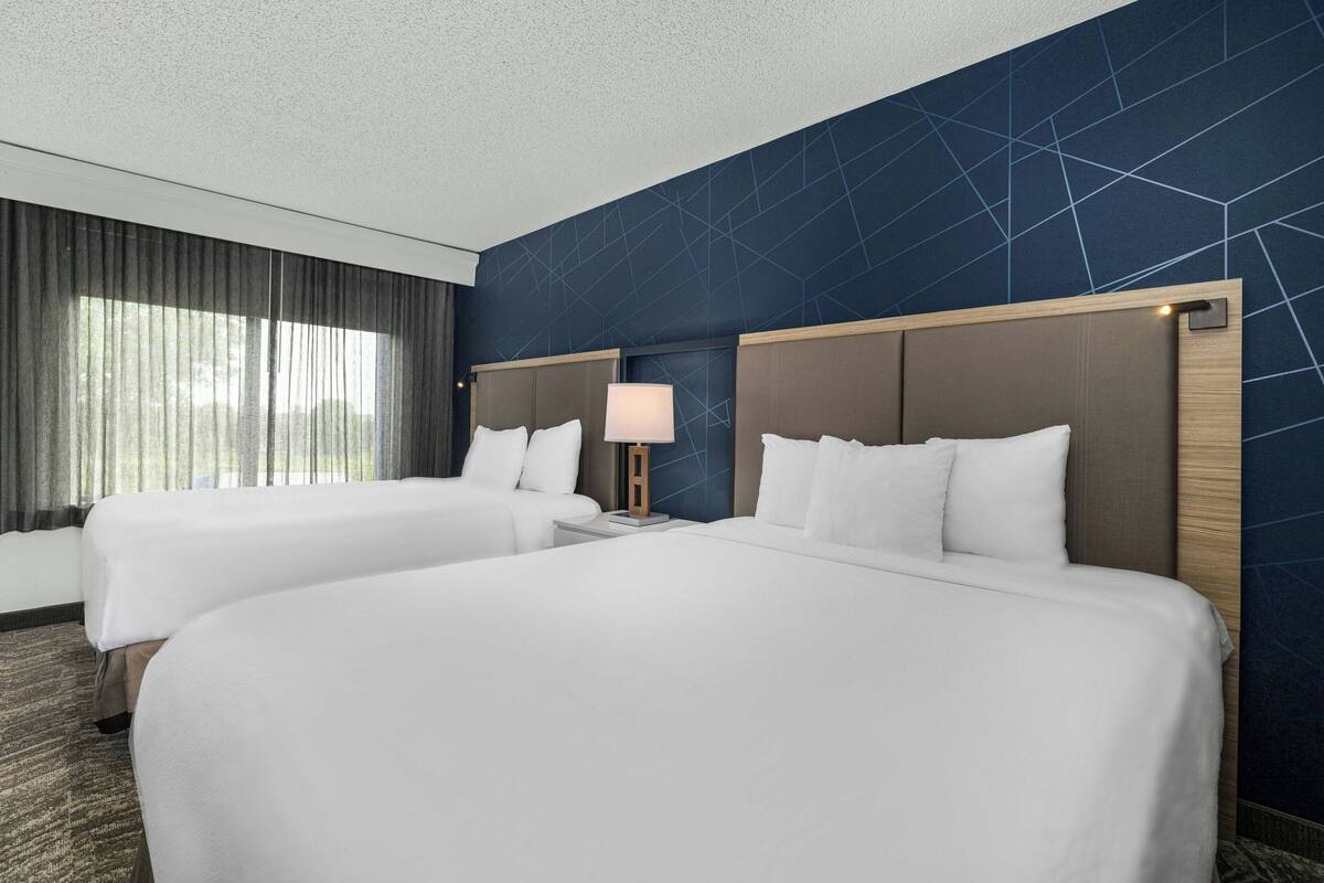 A hotel room with two beds, a nightstand with a lamp, and a large window with curtains.