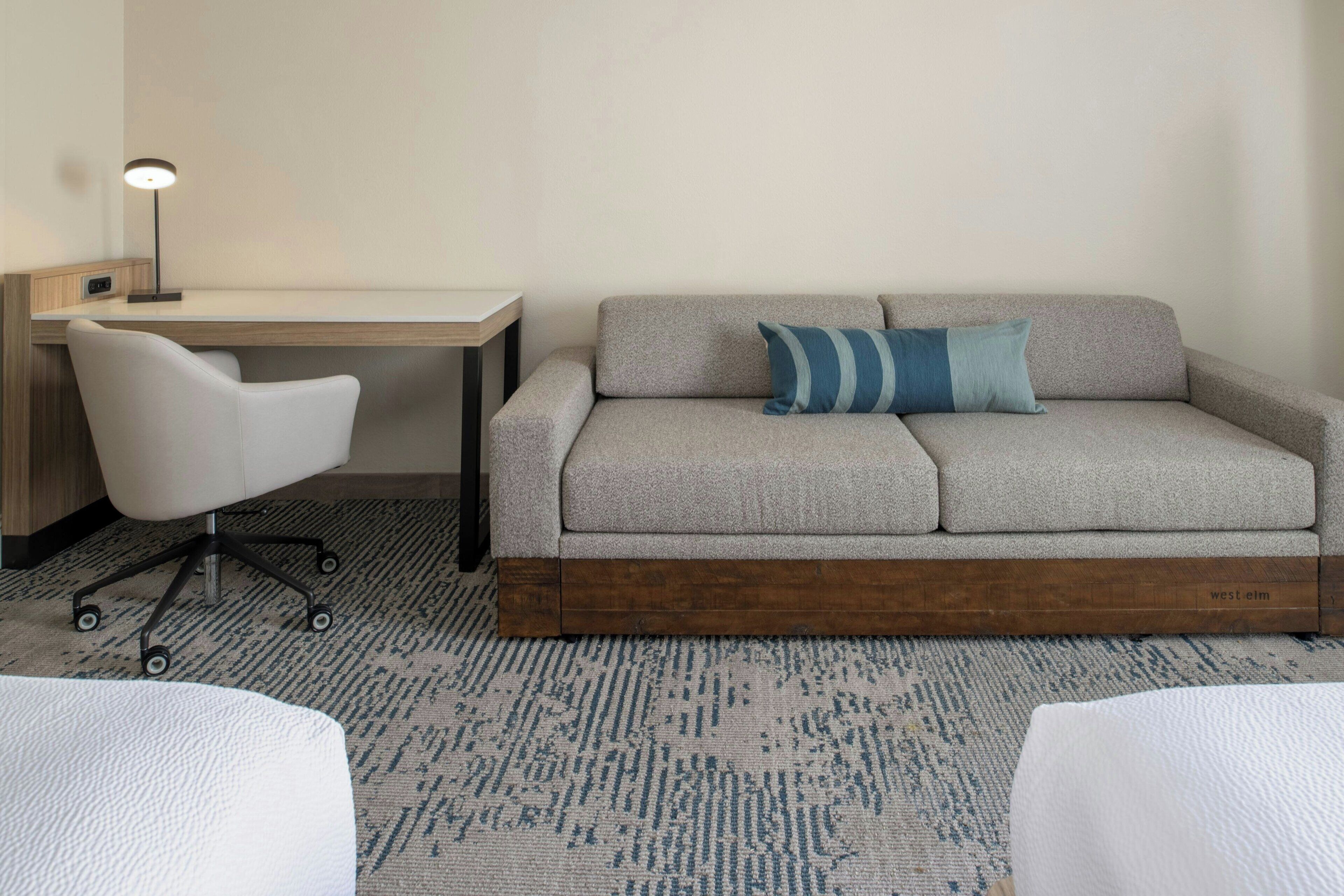 Une chambre d’hôtel avec un canapé gris, une chaise de bureau blanche, un bureau et une lampe.