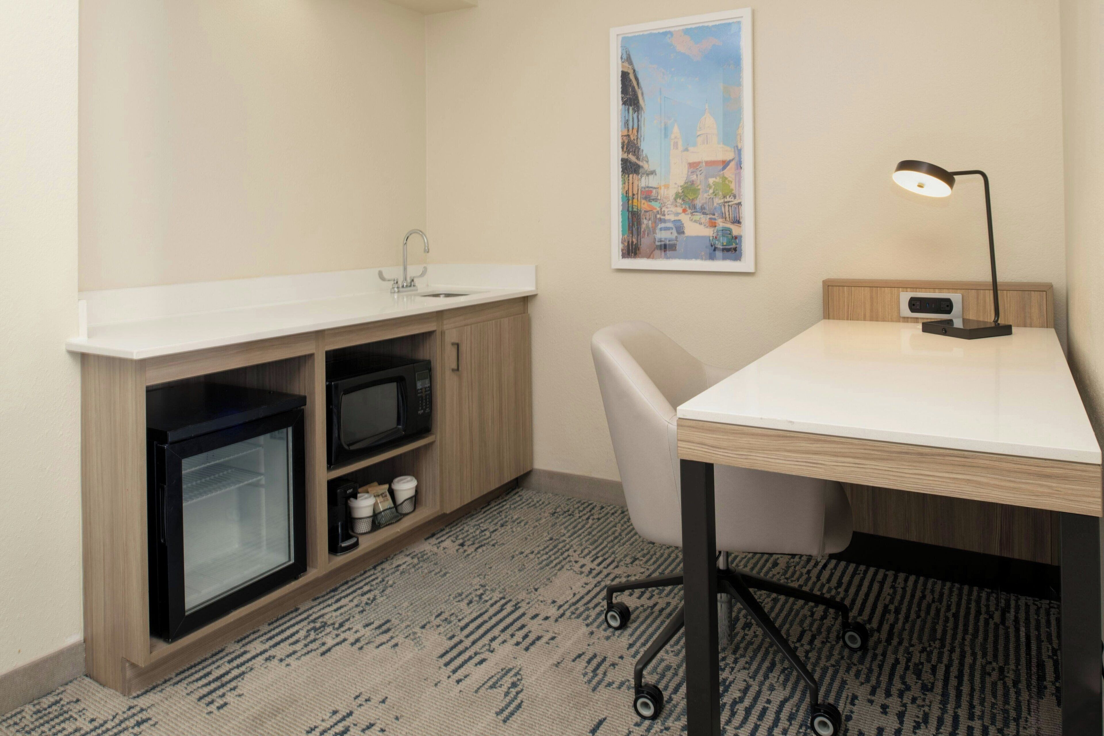 A compact hotel room with a kitchenette, a desk with a lamp, and a cityscape painting on the wall.