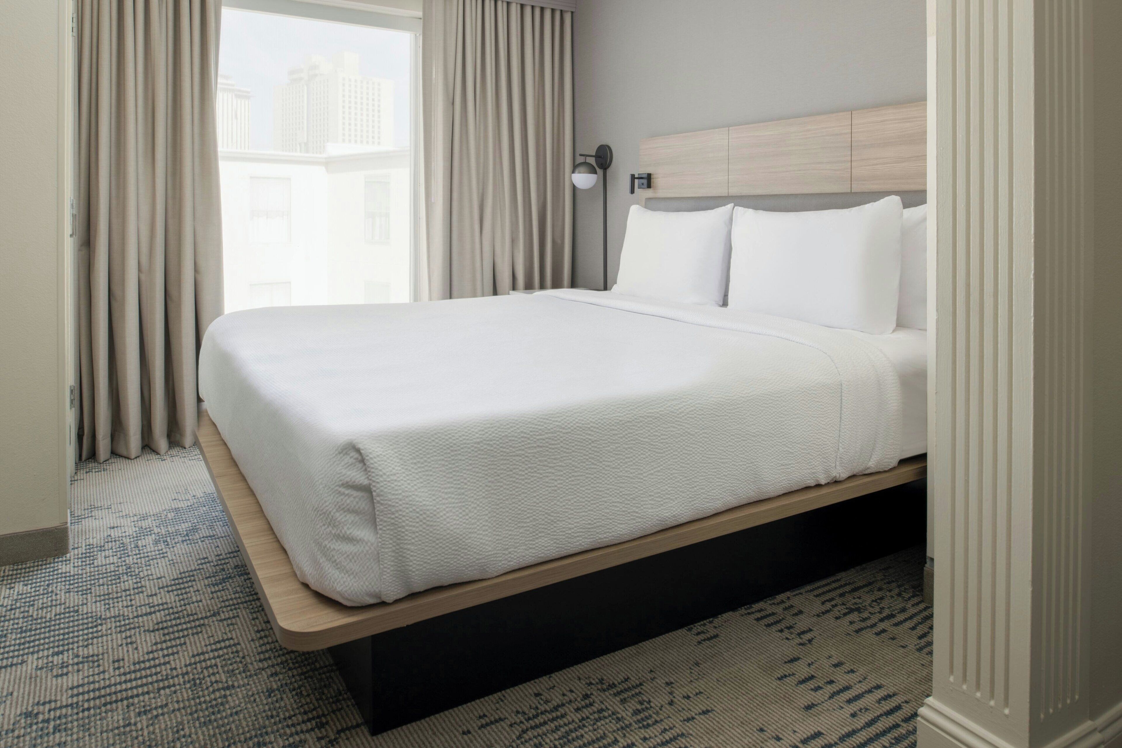 Habitación de hotel con una cama grande, cabecera, mesita de noche, lámpara y vista a edificios a través de la ventana.