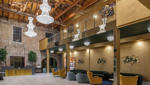 A spacious lobby with high ceilings, exposed brick walls, and modern chandeliers. There are multiple seating areas with plush sofas and armchairs.