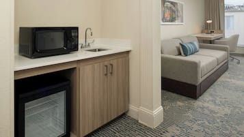 A compact hotel room with a kitchenette, a sofa, a small table, and a window with curtains.