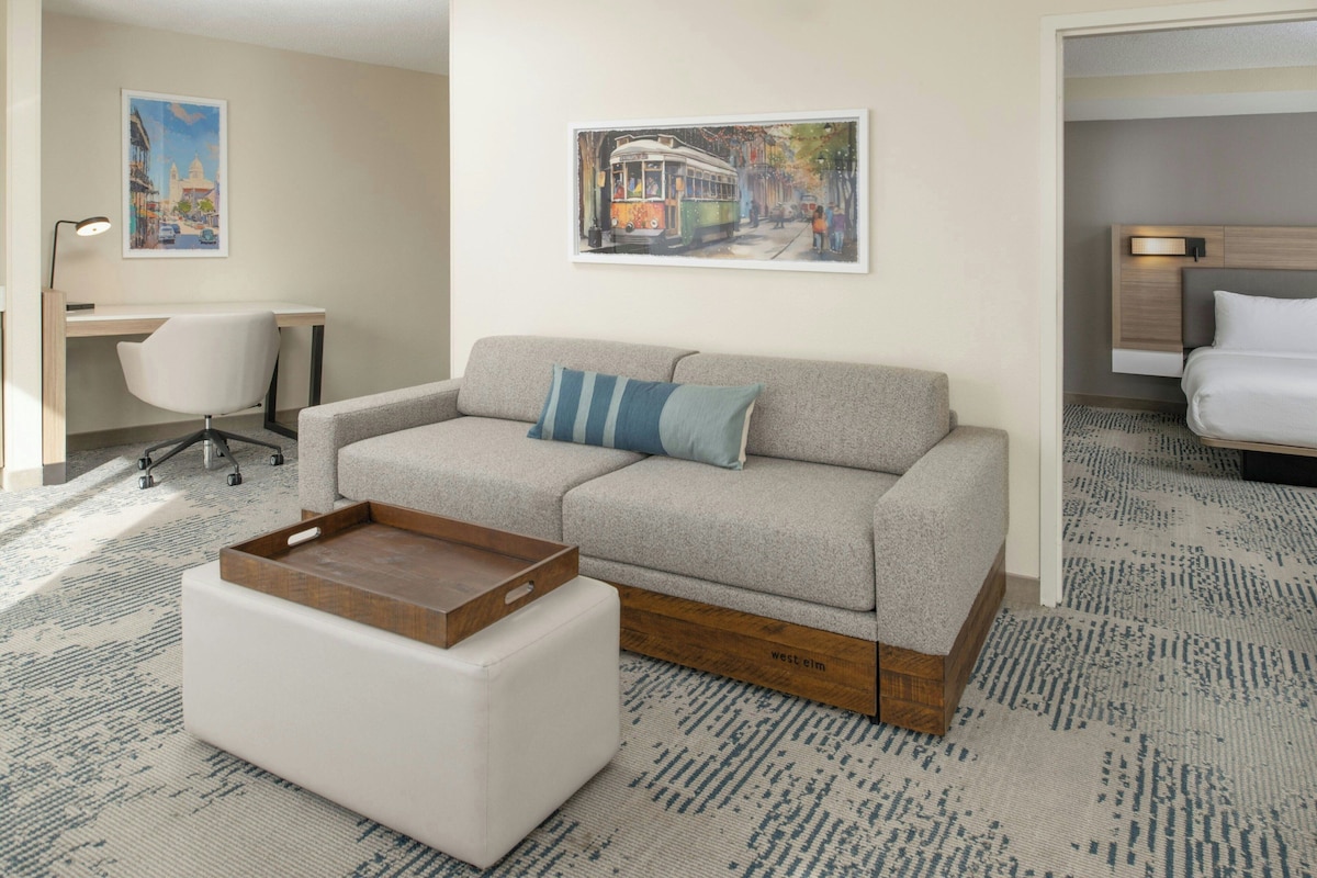 Una habitación de hotel moderna con un sofá gris, una mesita de madera, un escritorio con una silla y dos cuadros en la pared.