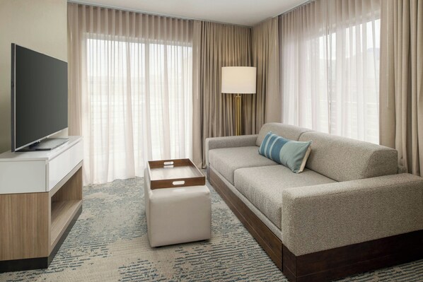 A modern hotel room with a flat-screen TV, a sofa with a blue pillow, a wooden side table, a white ottoman, and a patterned rug. - SpringHill Suites by Marriott New Orleans Warehouse Arts District (New Orleans)