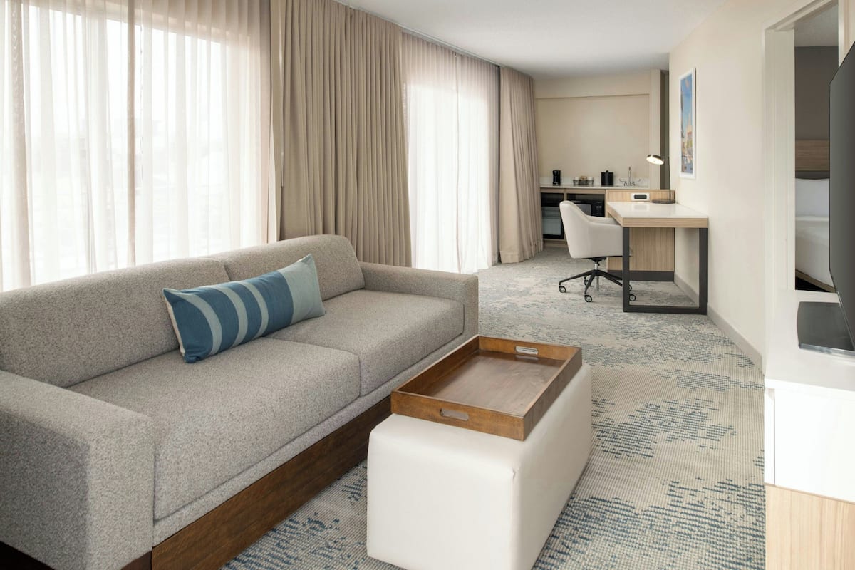 A modern hotel room with a sofa, a desk, a chair, and a large window with curtains.