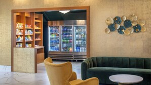 A lobby with a yellow armchair, a green sofa, a round metal table, a wooden shelving unit with snacks, and a refrigerator filled with beverages.