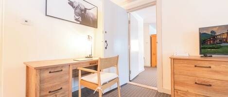 A hotel room with a wooden desk, chair, and dresser. There is a framed picture on the wall and a television on the dresser.