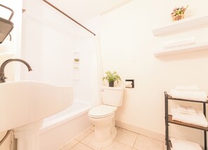 A bathroom with a white sink, toilet, and shower. There are shelves with toiletries and a plant.