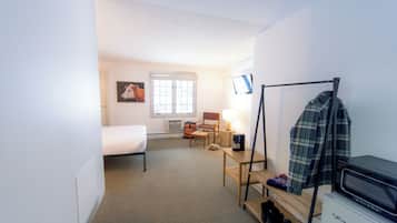 A compact hotel room with a single bed, a desk with a lamp, a chair, a television, and a coat rack.