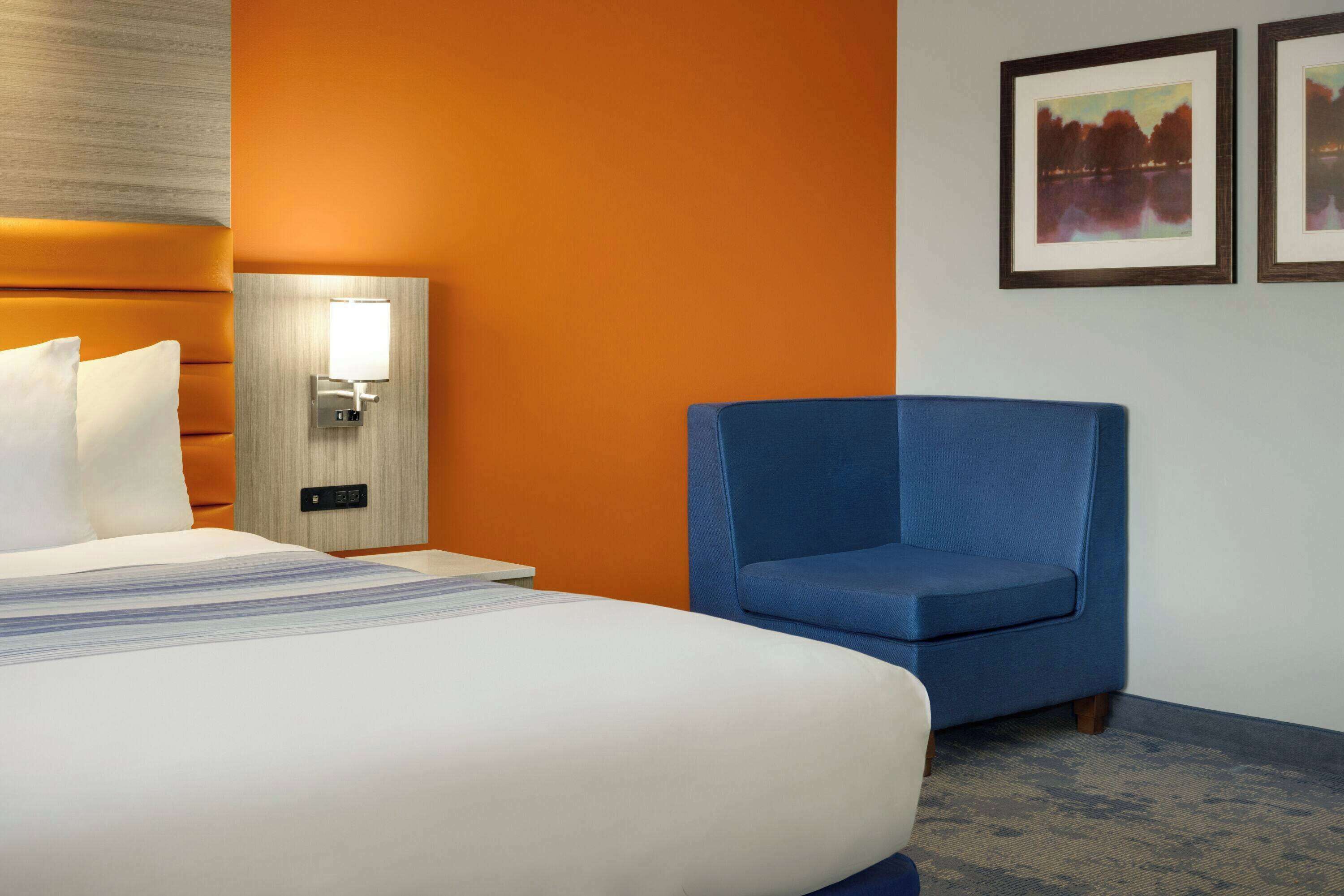 A hotel room with a bed, a blue armchair, a wall-mounted lamp, and two framed pictures on the wall.