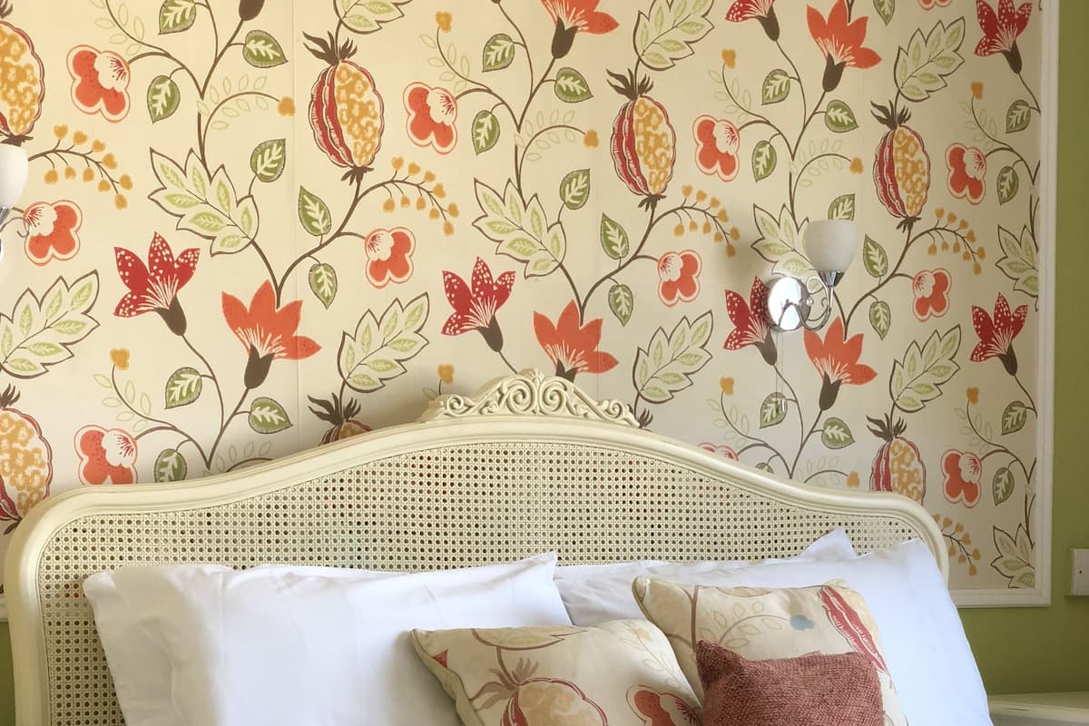 A bed with white bedding and multiple pillows, a woven headboard, and a floral wallpaper pattern.