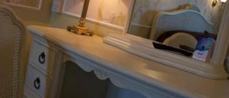 A hotel room with a white vanity, a mirror, a lamp, a bed with a headboard, and a plaid chair.