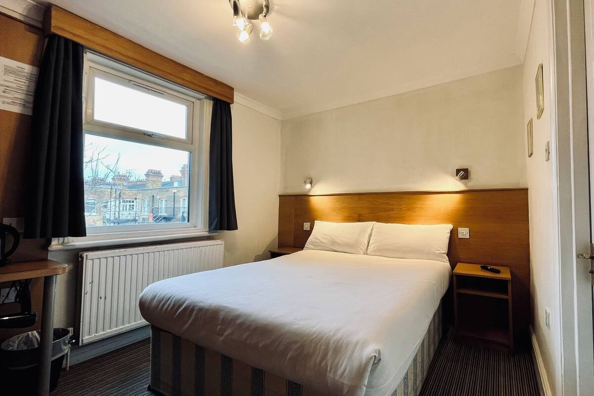 A hotel room with a bed, a window, a radiator, a bedside table, and a nightstand.