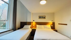A hotel room with two single beds, a nightstand with a lamp, a framed picture on the wall, and a window with a view of trees. - West Cromwell Hotel (London)