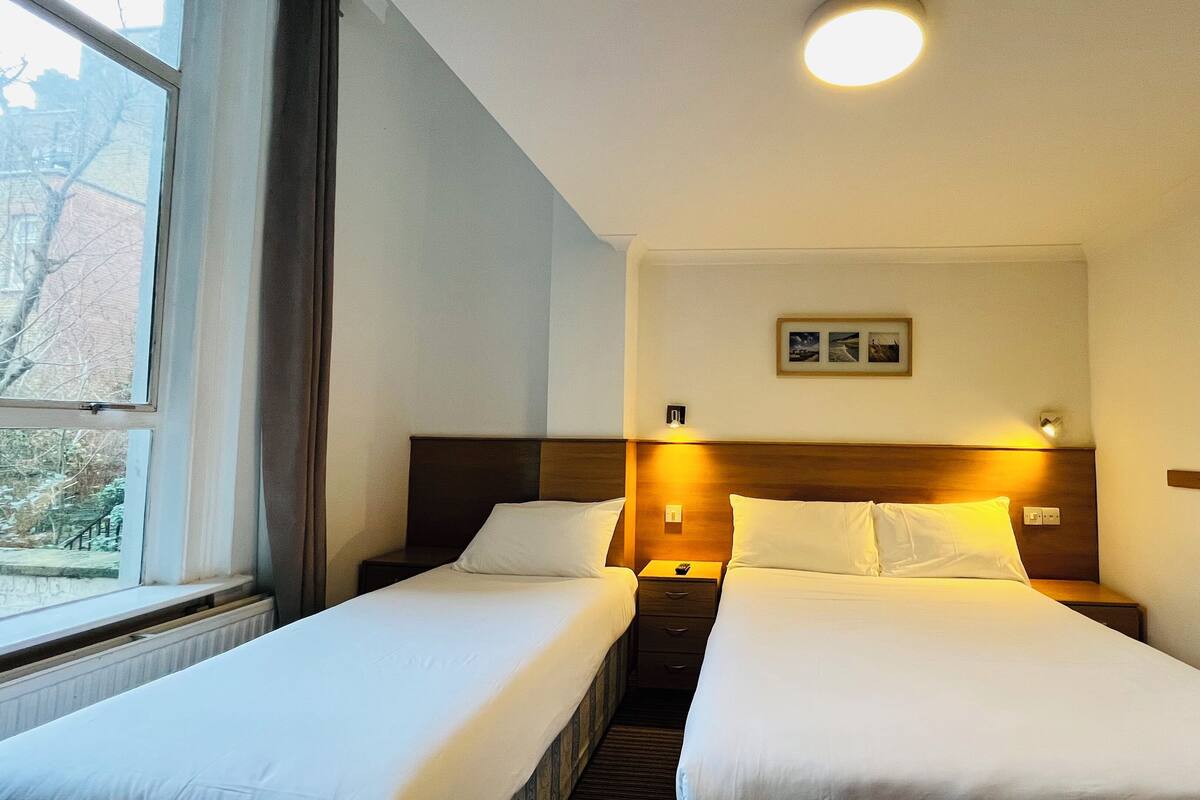 A hotel room with two single beds, a nightstand with a lamp, a framed picture on the wall, and a window with a view of trees.