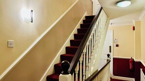 A staircase with red carpeted steps and white balusters, leading to a door with a fire extinguisher mounted on the wall. - West Cromwell Hotel (London)