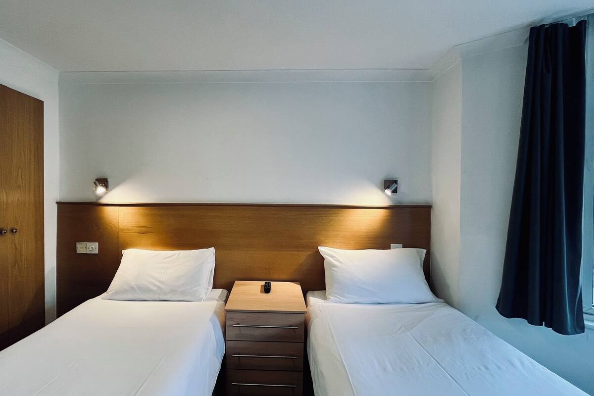 A hotel room with two single beds, a wooden headboard, bedside lamps, a nightstand, and a window with curtains.