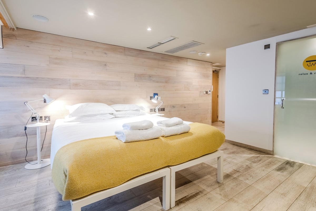 Una habitación de hotel moderna con suelo de madera, cama con ropa blanca, manta amarilla, mesita de noche con lámpara y puerta de vidrio con un logotipo.