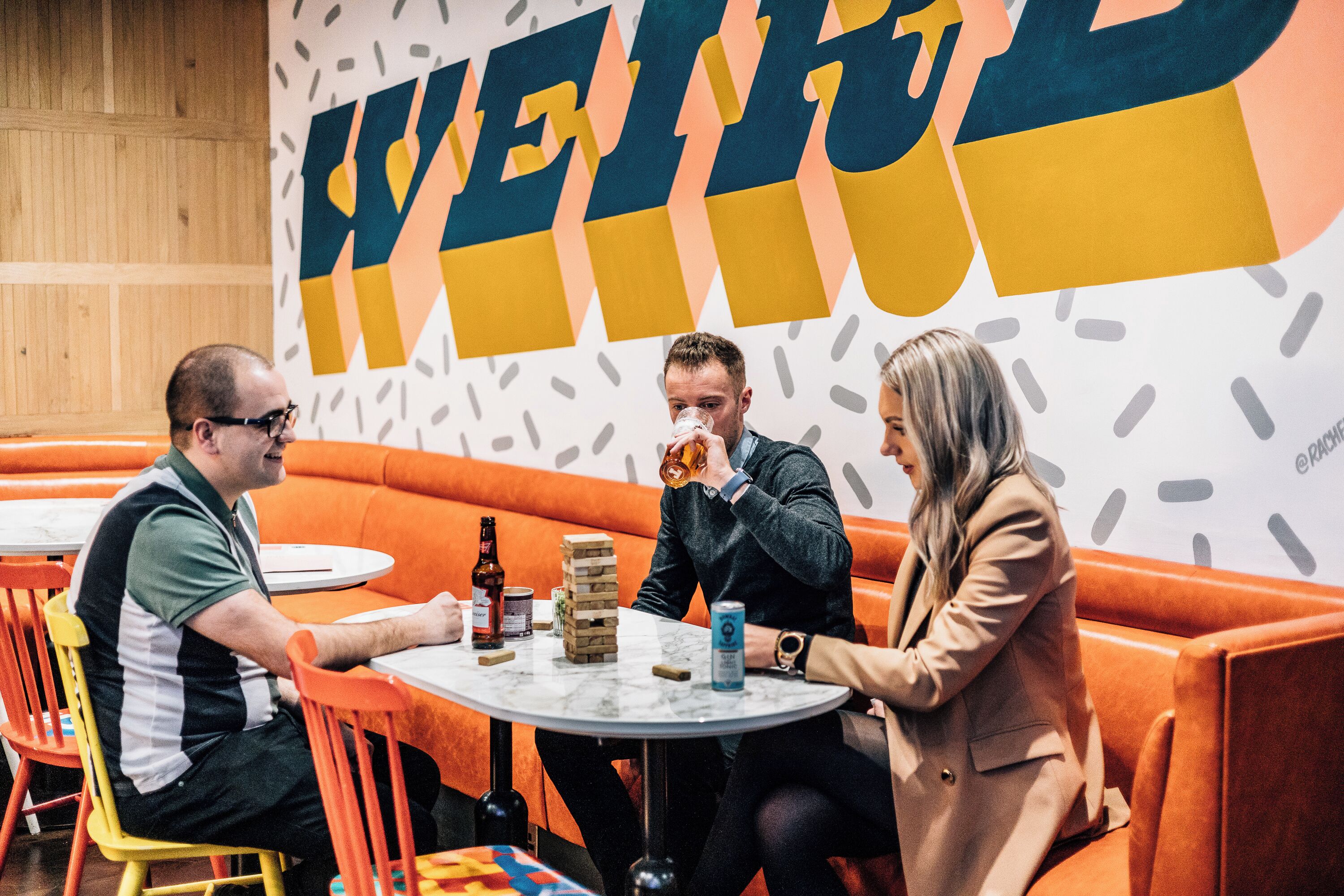 A group of people seated at a table in a brightly lit indoor setting with orange seating and a large wall with stylized text.