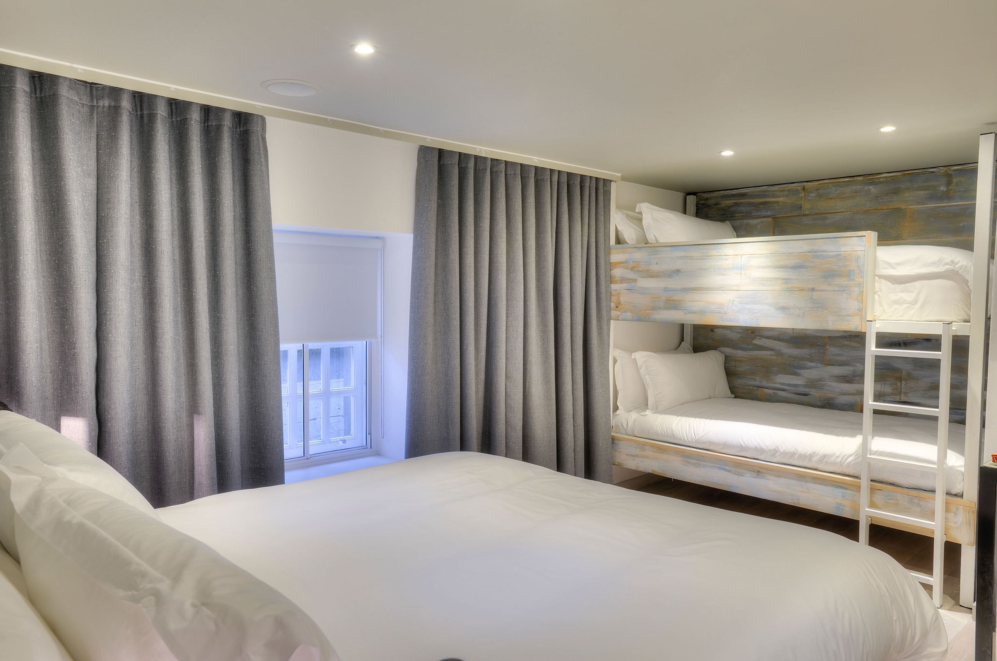 A bedroom with a bunk bed, a large bed, grey curtains, and a view of a balcony.