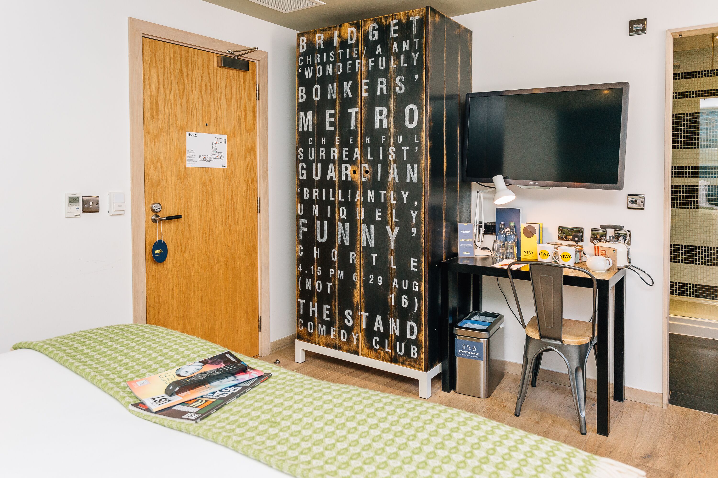 A room with a bed, a desk with a computer and a TV, a wardrobe with words on it, and a door with a sign.