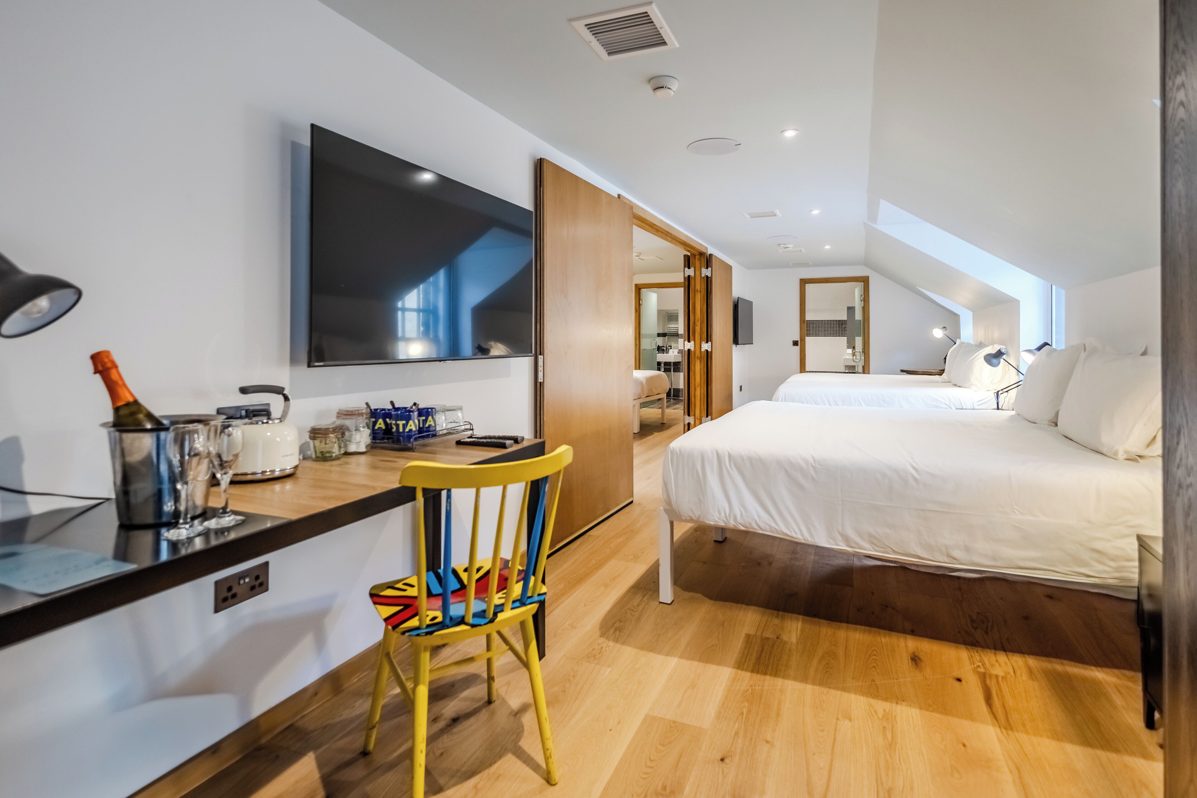 A modern hotel room with a bed, a desk with a chair, a TV, and a large window.
