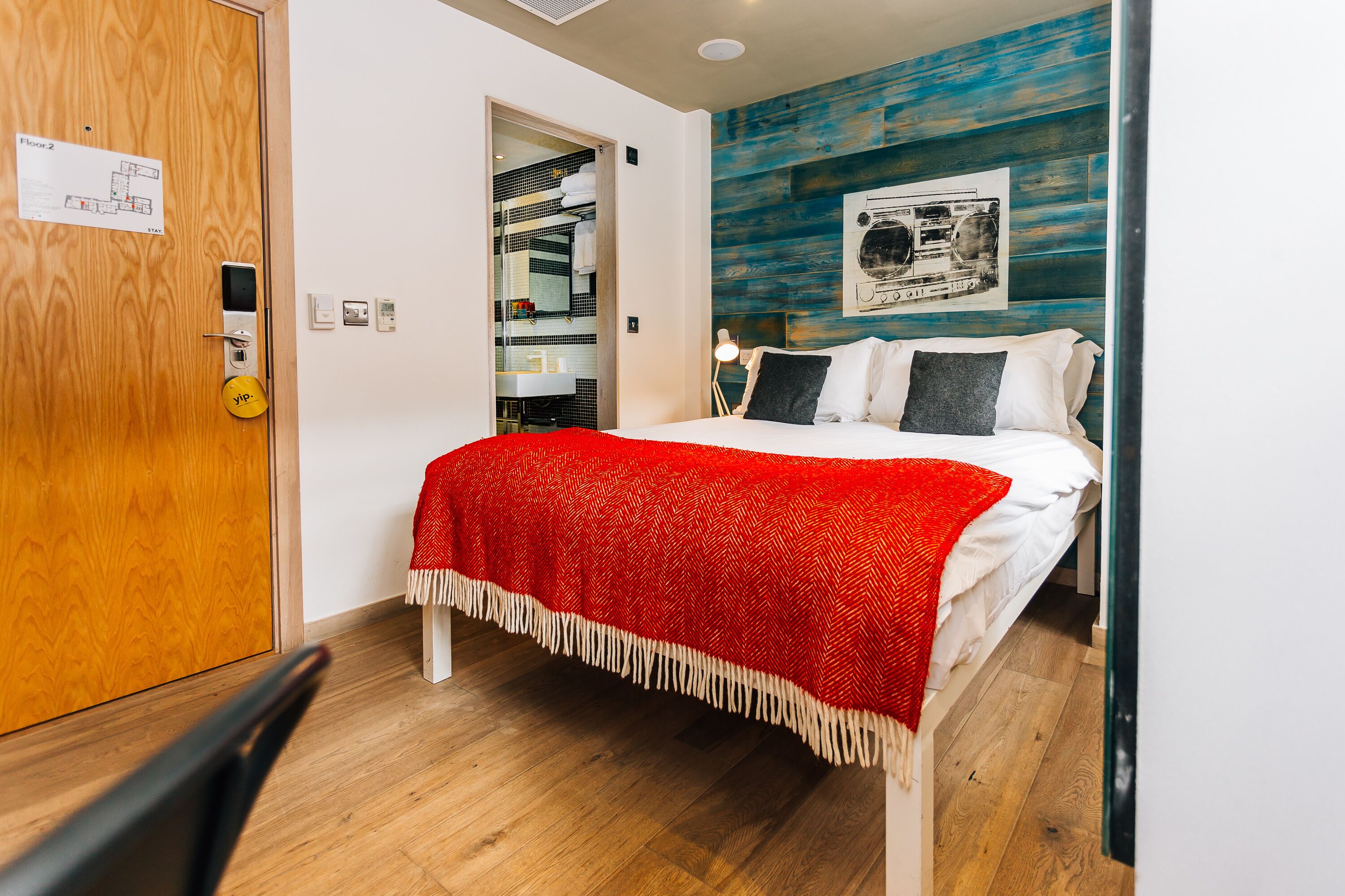 A hotel room with a bed, a red throw blanket, a wooden floor, a door with a keypad lock, and a wall with a blue painted section.