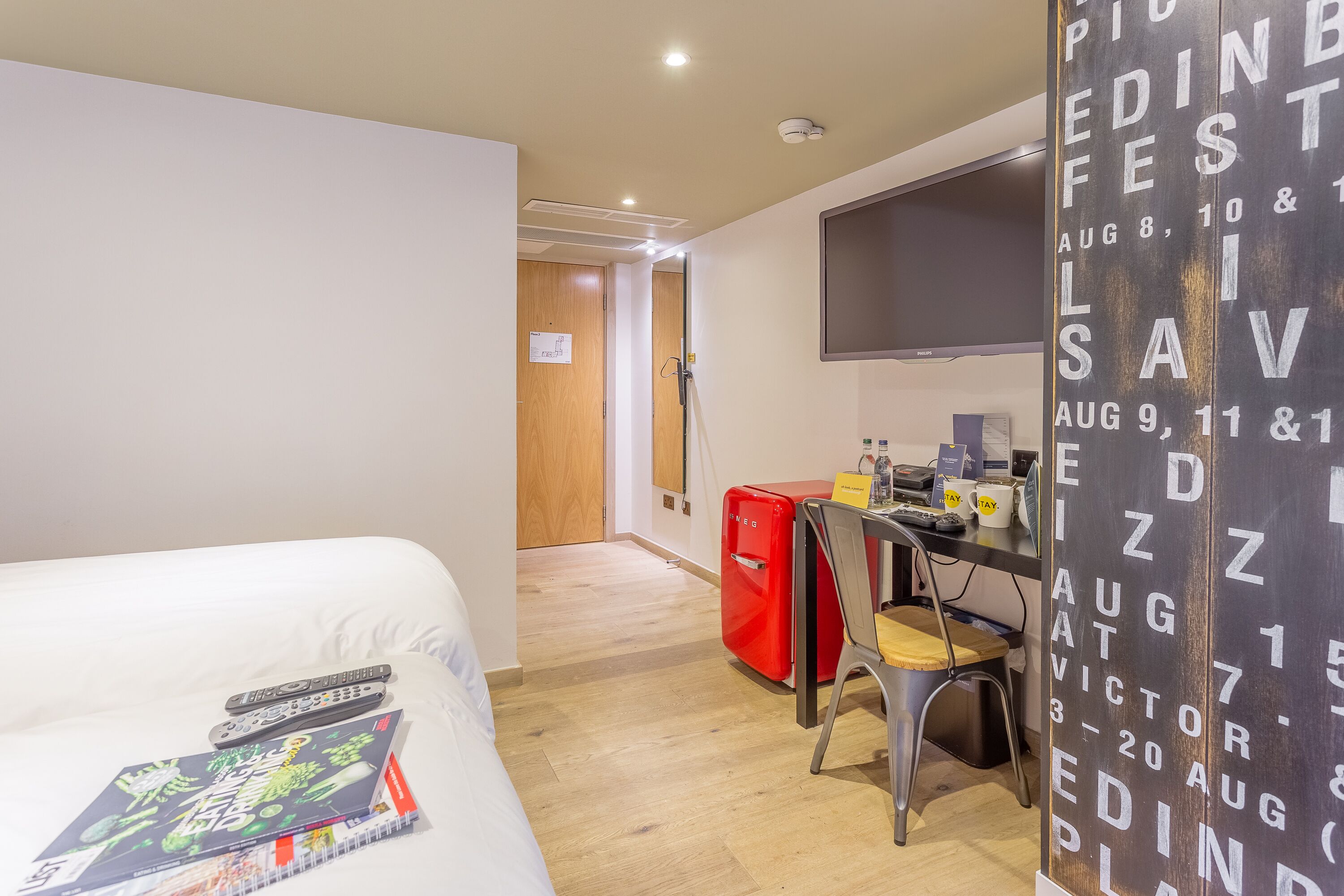 A hotel room with a bed, a desk with a chair, a red suitcase, and a wall-mounted TV.