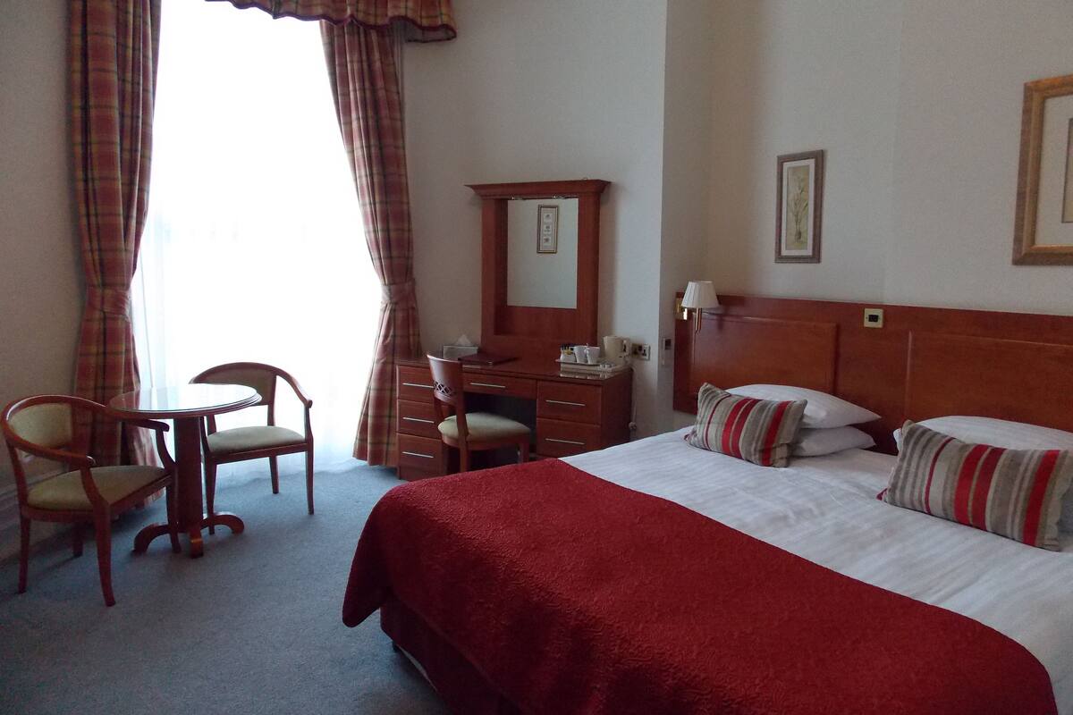 A hotel room with a bed, a desk, two chairs, a dresser, and a window with curtains.