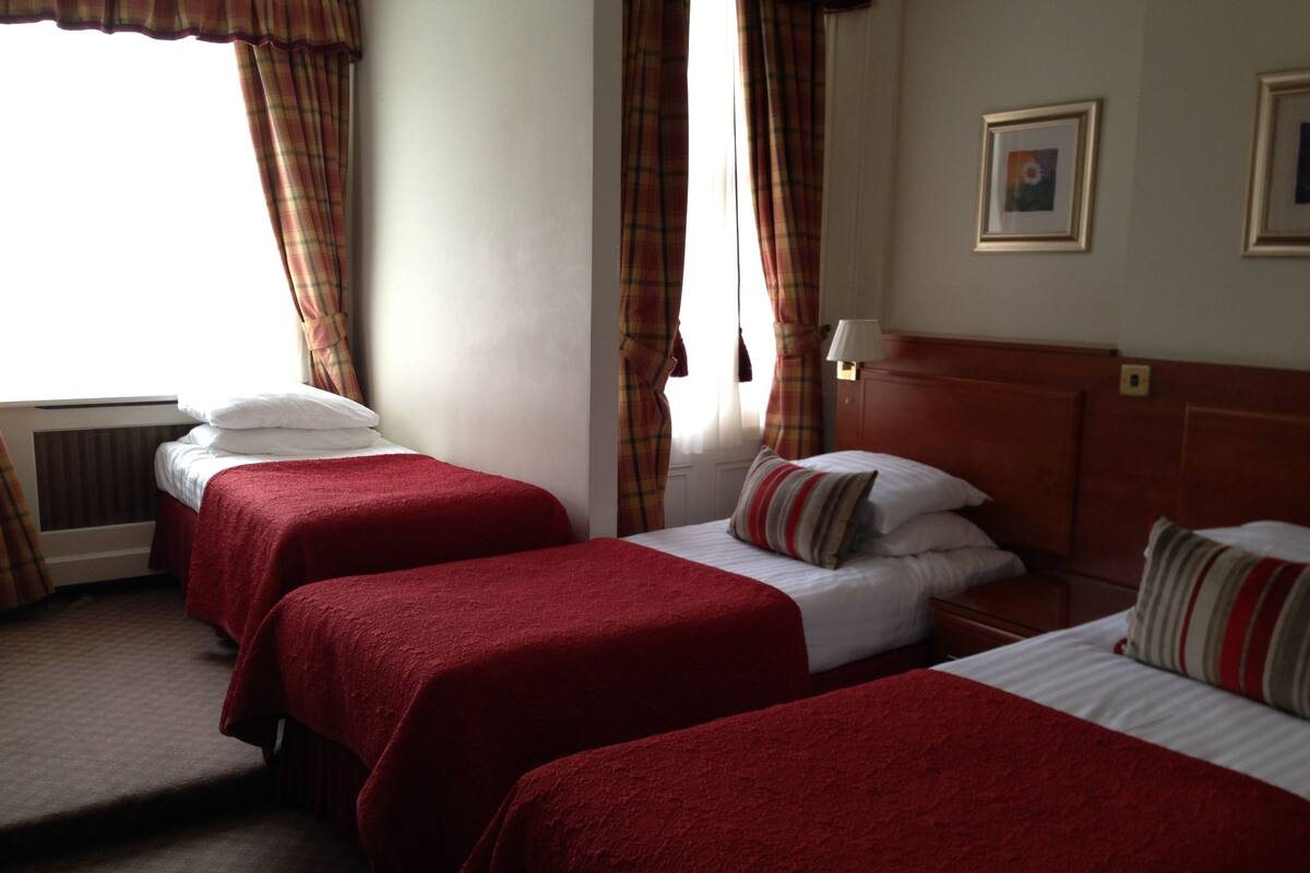 A hotel room with two beds, each with red bedding and white pillows. There is a window with curtains, a radiator, and a framed picture on the wall.