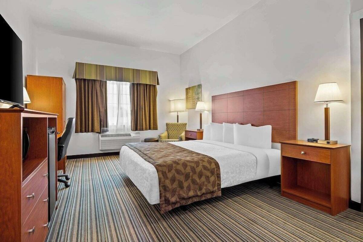 A hotel room with a large bed, a desk with a computer, a TV, and a window with curtains.