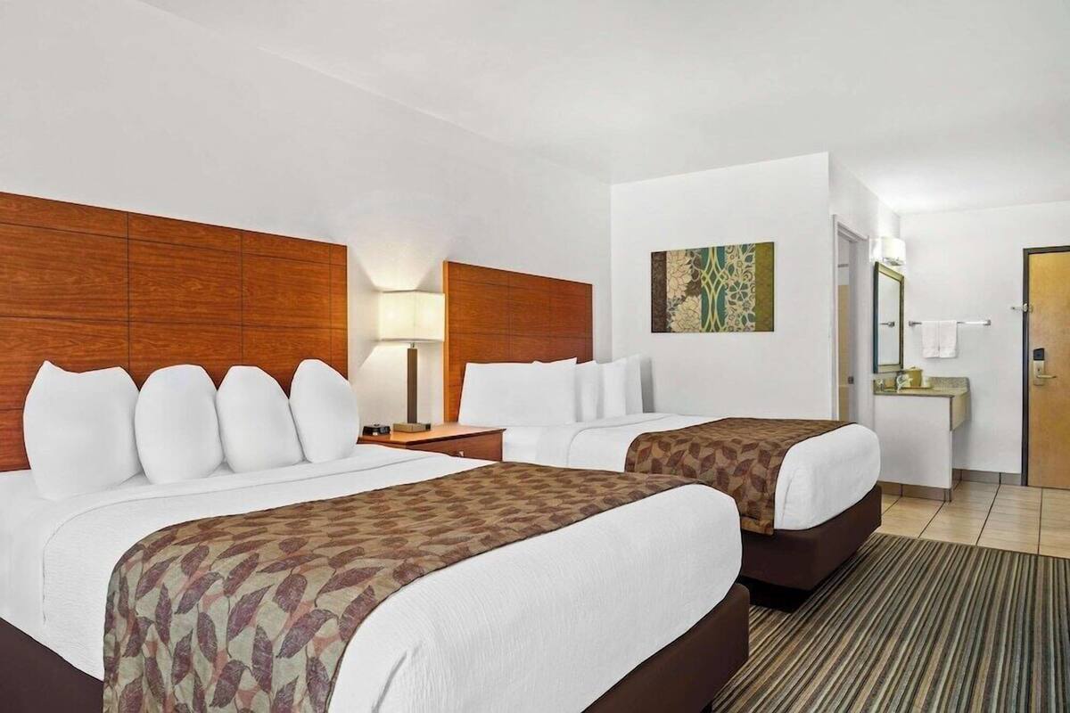A hotel room with two beds, a wooden headboard, a nightstand, a lamp, a painting, a bathroom with a mirror, and a door.