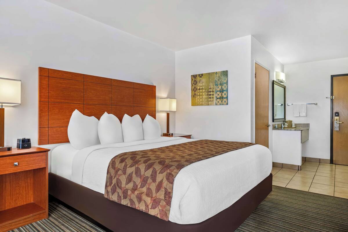 A hotel room with a large bed, a wooden headboard, a bedside table with a lamp, a small desk with a chair, a bathroom with a sink and mirror, and a painting on the wall.