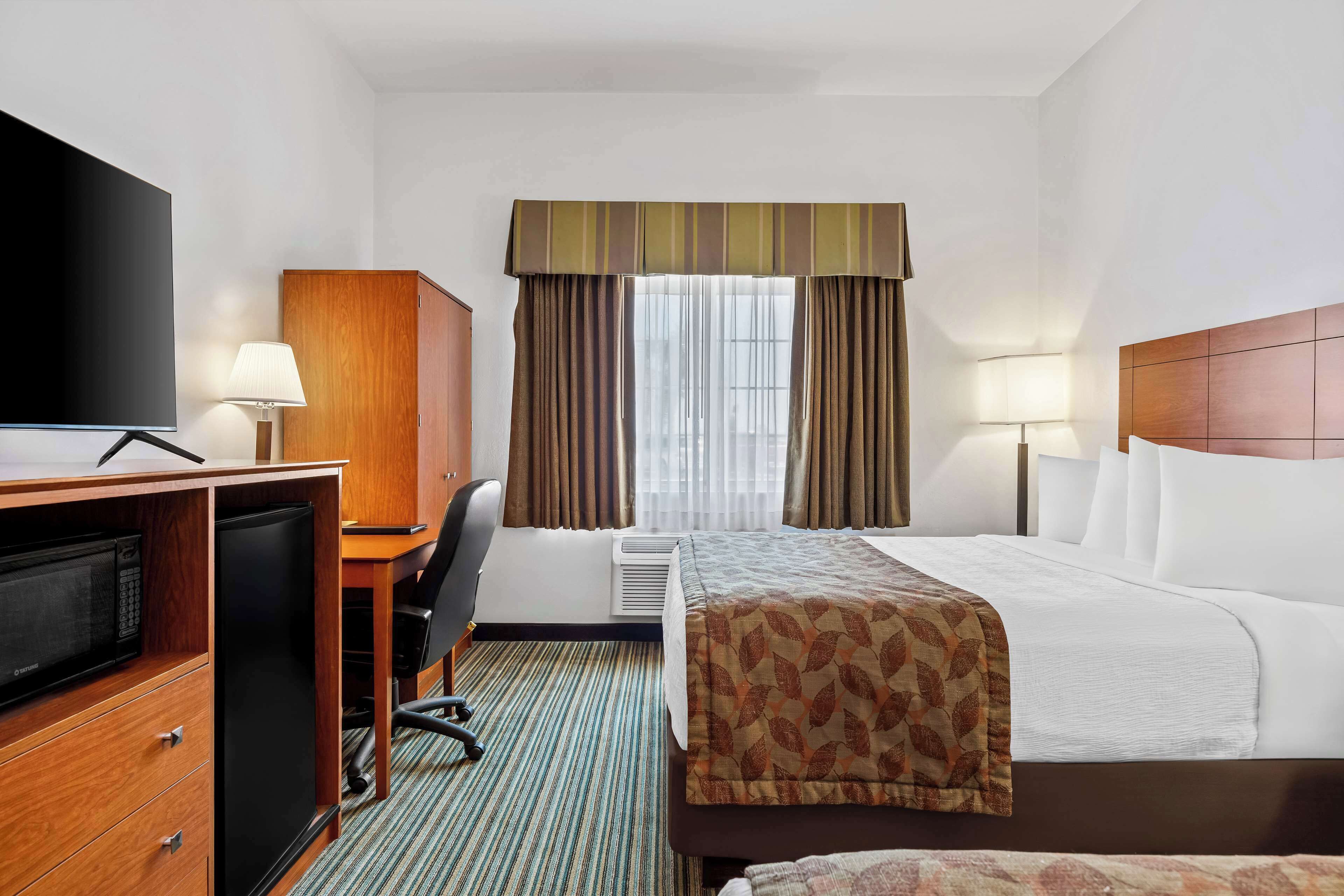 Quarto de hotel com cama, escrivaninha com televisão, frigorífico, cadeira e janela com cortinas.
