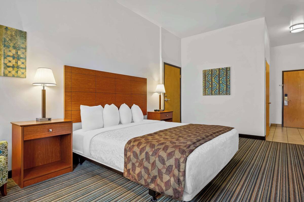 A hotel room with a large bed, two bedside tables with lamps, a wooden headboard, a patterned throw blanket, and a carpeted floor.