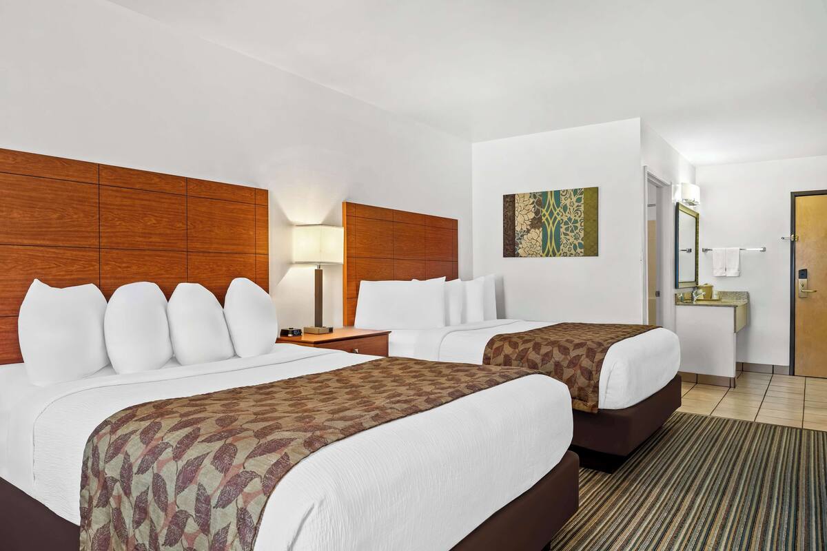 A hotel room with two beds, a wooden headboard, a bedside table with a lamp, a bathroom with a sink and mirror, and a patterned rug.
