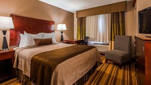 A hotel room with a large bed, a desk, a chair, a television, and a window with curtains. - Old Mill Inn (Omaha)