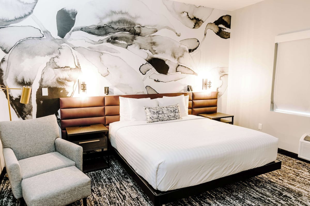 Une chambre d’hôtel avec un grand lit, un fauteuil gris, une œuvre d’art moderne et abstraite accrochée au mur, et deux lampes de chevet.