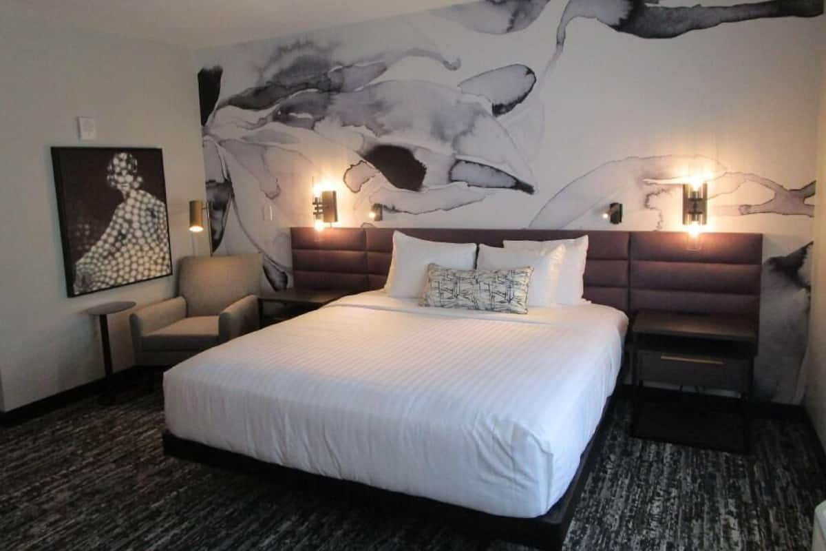 Une chambre d’hôtel avec un grand lit, deux tables de chevet, une chaise, une œuvre d  ’art murale et une fresque murale moderne et abstraite.