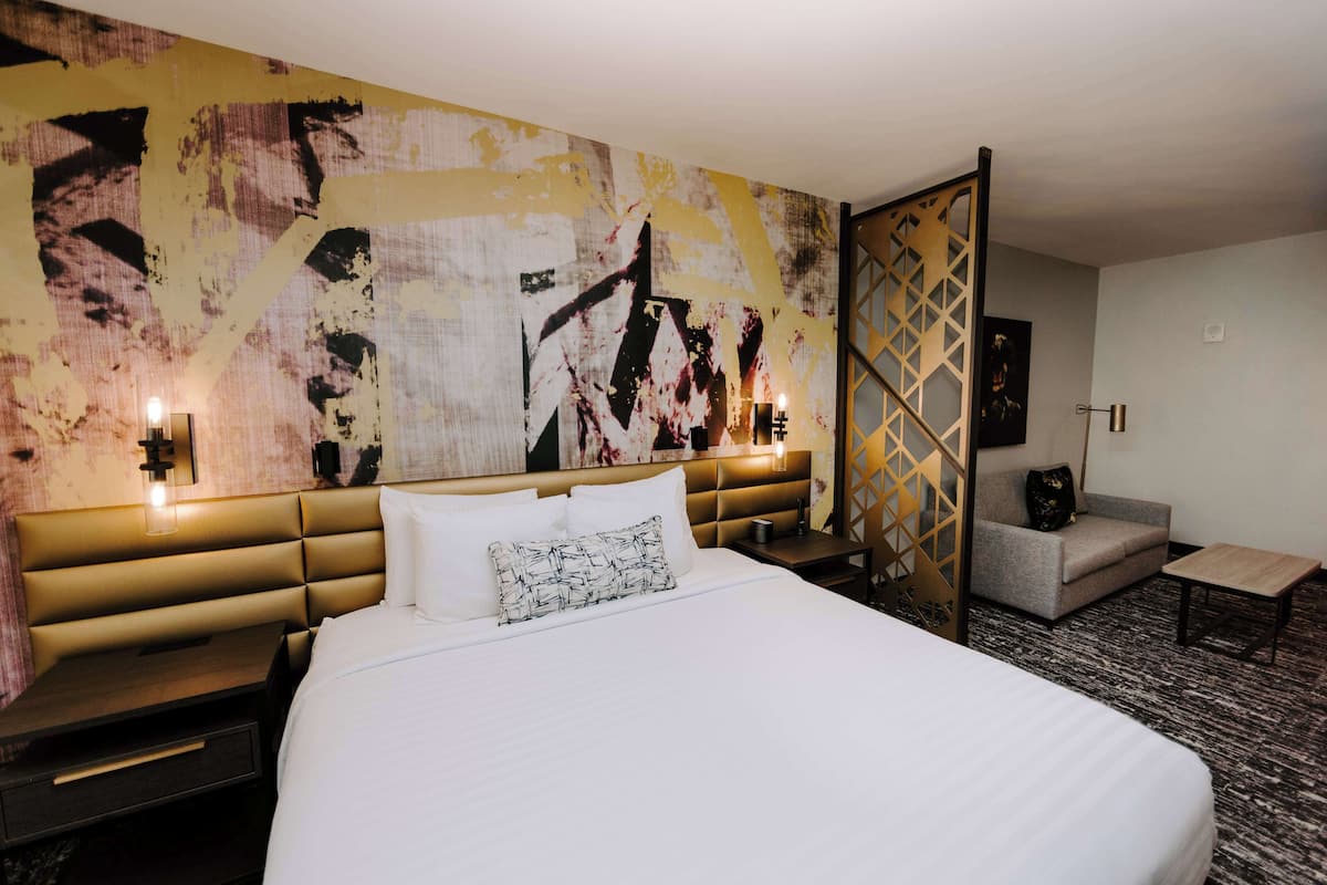 Une chambre d’hôtel avec un grand lit, une tête de lit dotée d’un éclairage intégré, un mur orné d’un tableau abstrait, un canapé et une petite table.