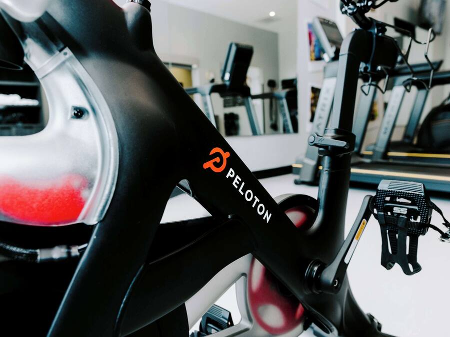 A Peloton stationary bike in a gym with exercise equipment in the background.