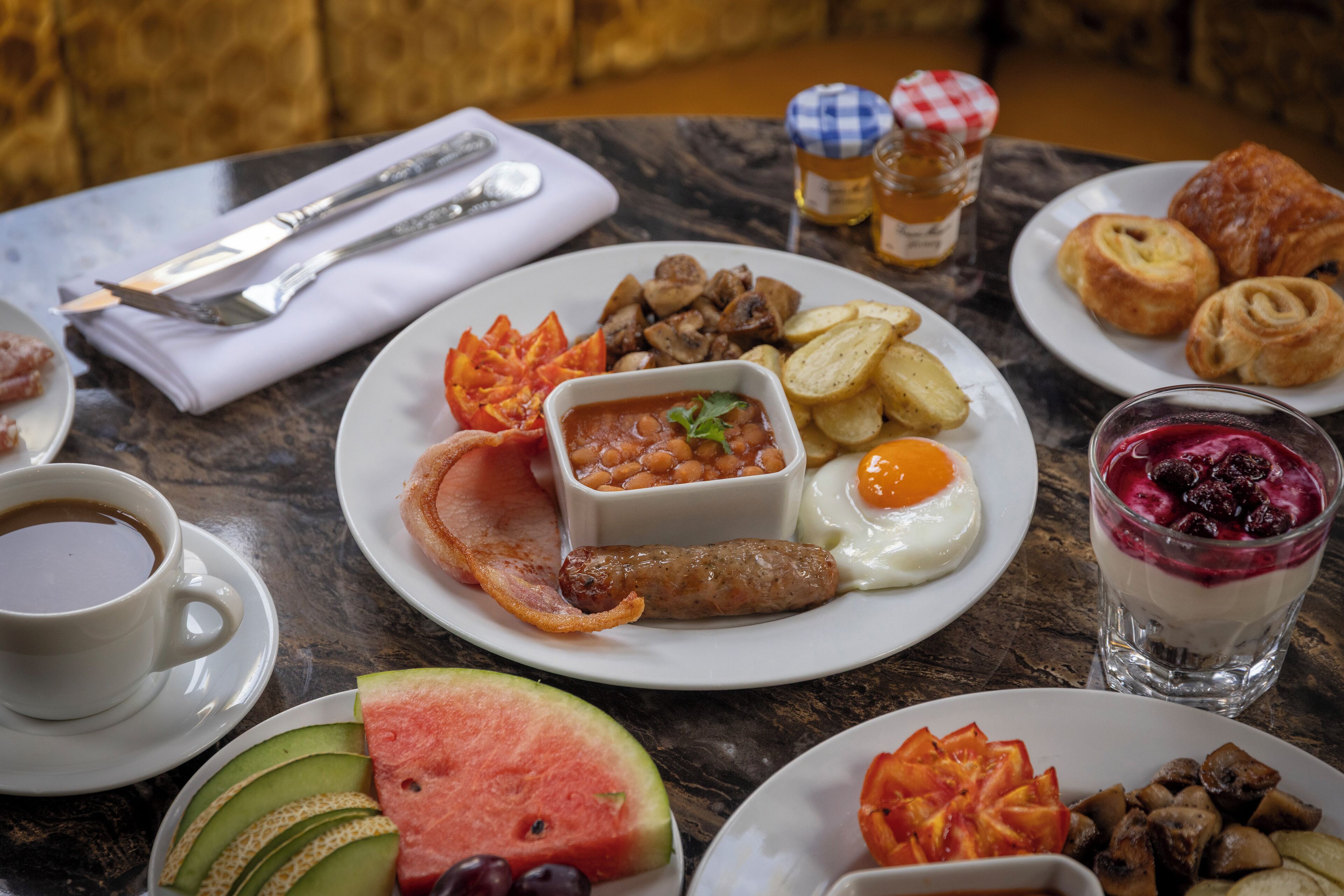 A breakfast table with plates of food, a cup of coffee, and a glass of yogurt parfait.