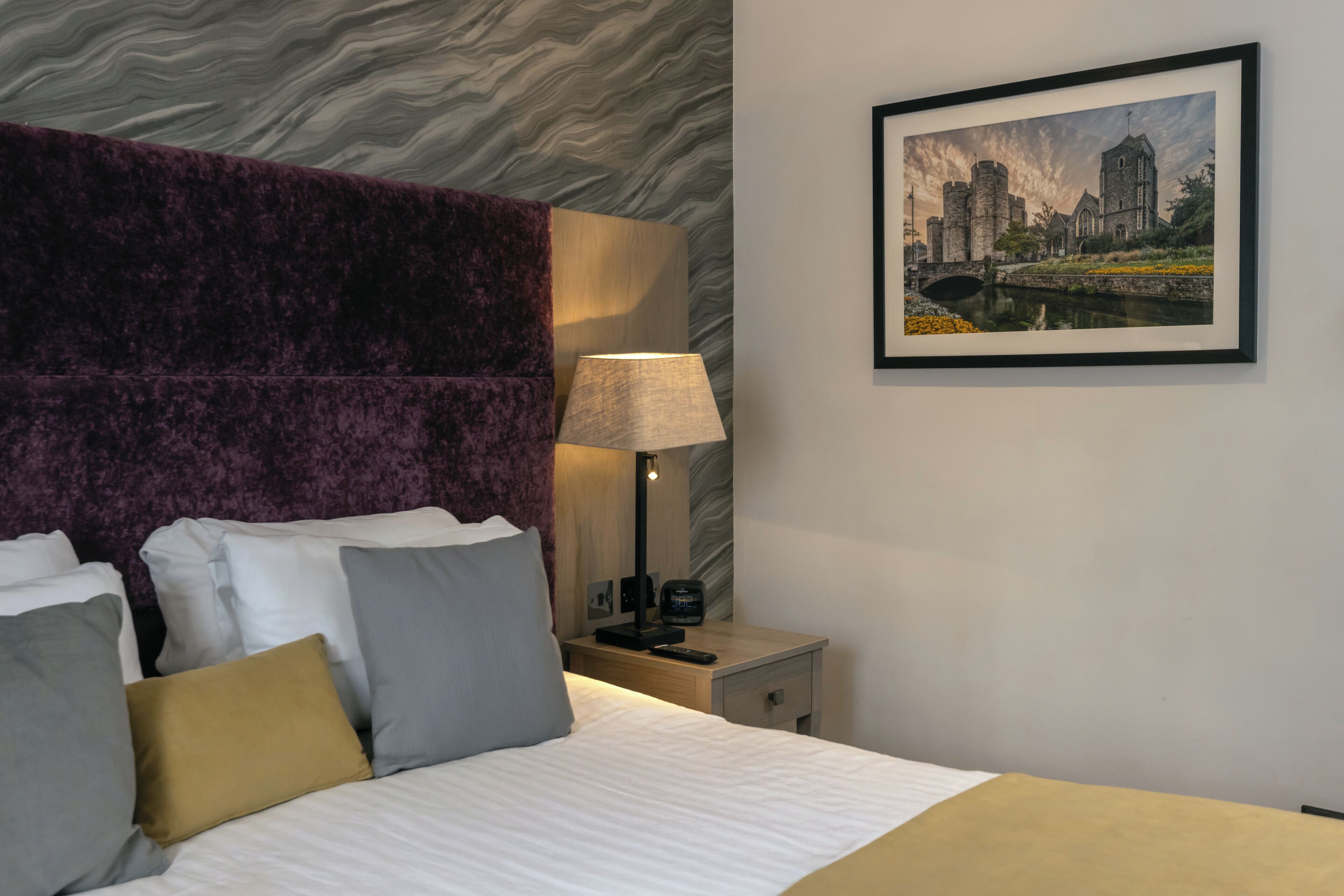 A hotel room with a large bed, a headboard, a bedside table with a lamp, a framed picture on the wall, and a nightstand.